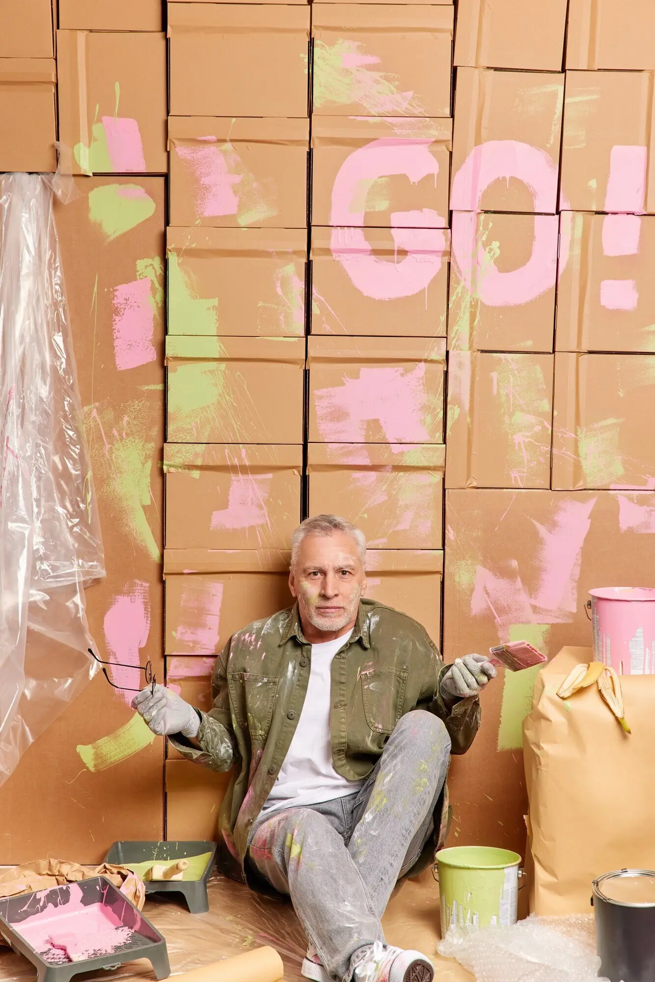 Innenaufnahme eines grauhaarigen Mannes, der auf dem Boden sitzt, zögernd mit den Schultern zuckt, die Brille abnimmt und legere Kleidung trägt, umgeben von einem Farbeimer und Reparaturwerkzeugen. Bau, Umbau oder Renovierung.